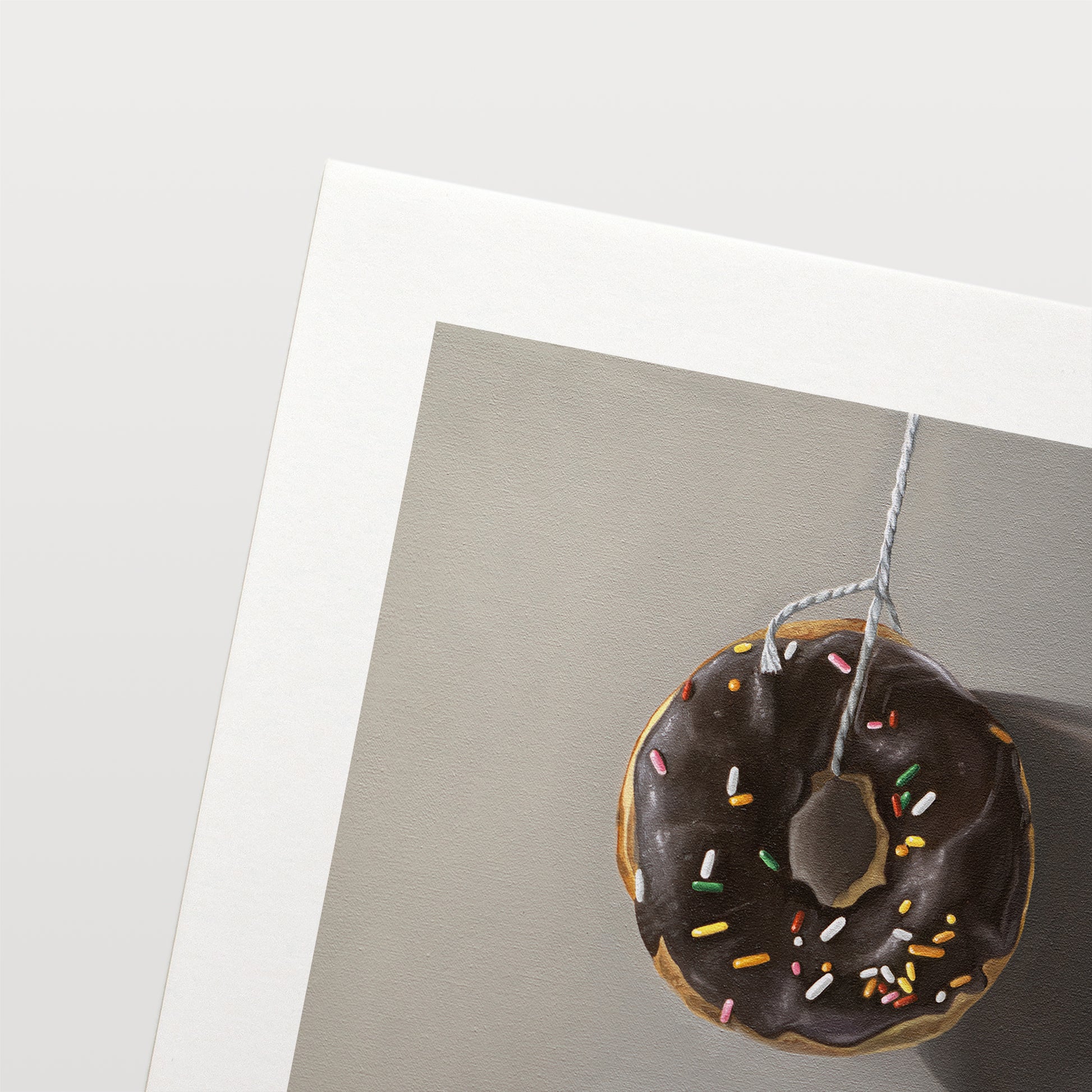 This artwork features a chocolate sprinkled doughnut suspended by a string over a white porcelain cup.