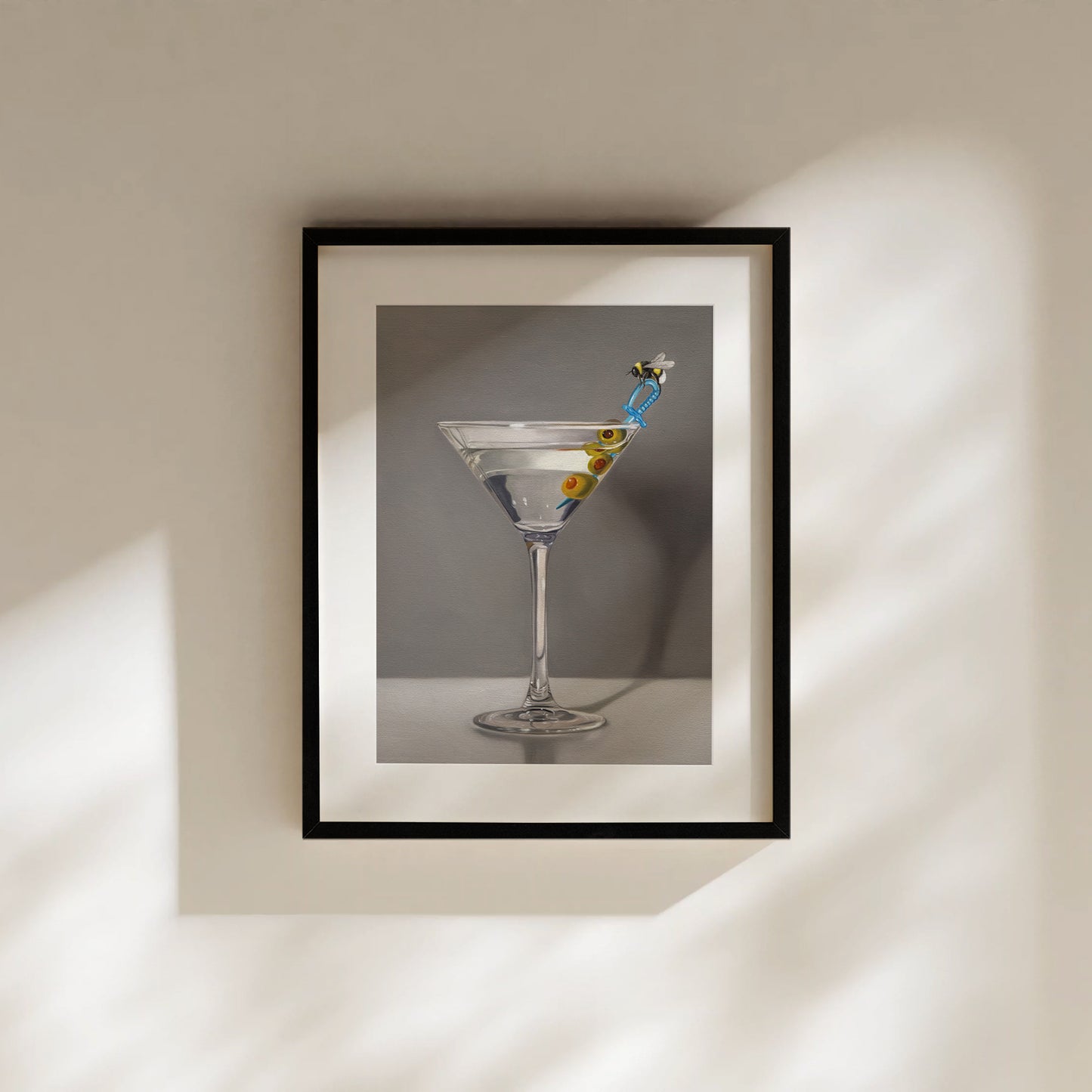 Framed fine art print of a martini with olives and a bumble bee stirrer, on a light wall.
