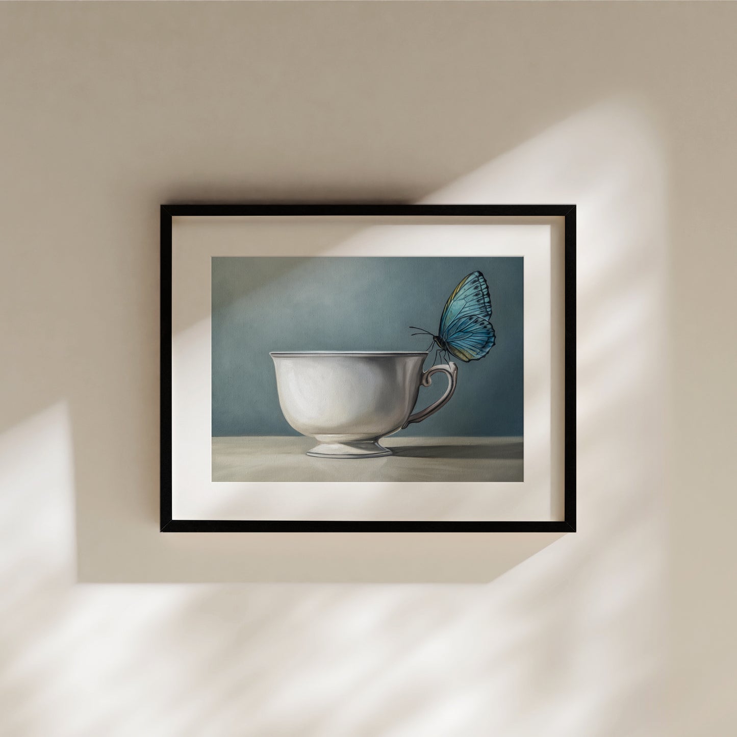 Framed fine art print of a blue butterfly perched on a white teacup against a muted blue background.