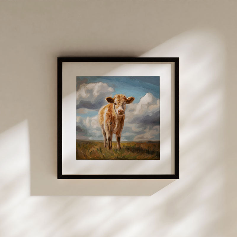 Curious Cow fine art print in a black frame, featuring a brown cow in a grassy field under a cloudy blue sky.