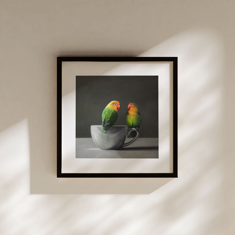 Fine art print of two green lovebirds perched on a grey porcelain cup, framed in black against a neutral wall.
