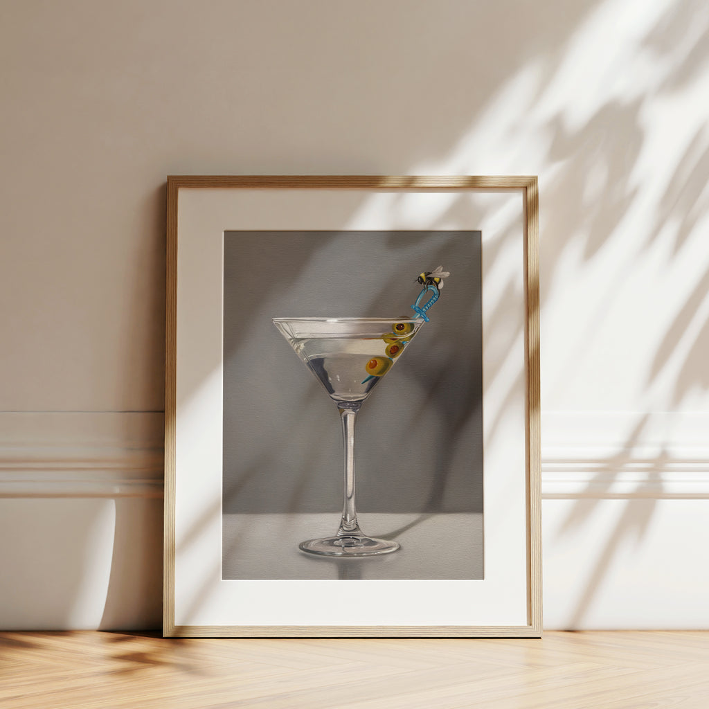 This artwork features a martini with a bumble bee perched on a blue plastic cocktail sword with a trio of green olives and a neutral, minimalistic backdrop.