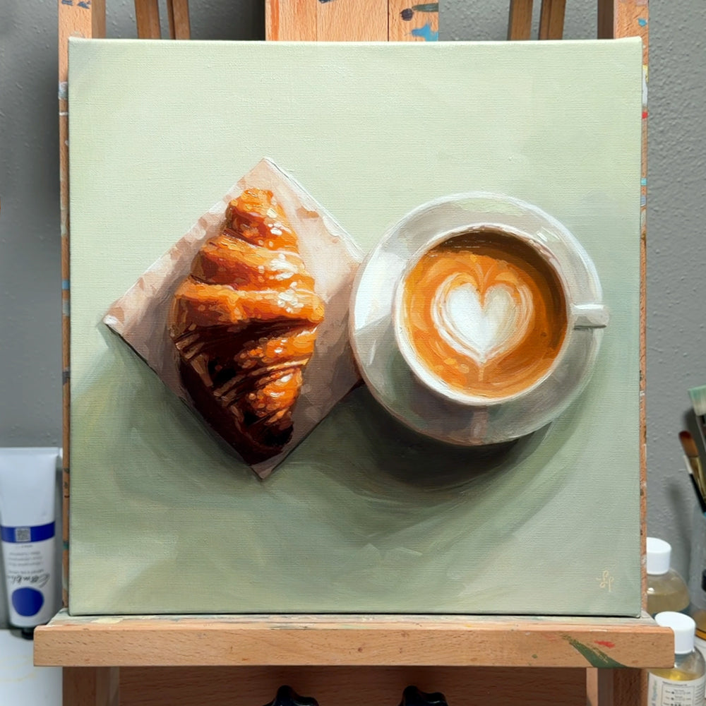 A square canvas painting on an easel depicting a croissant on parchment paper and a cup of latte coffee with a heart-shaped latte art design, set against a muted sage green background.