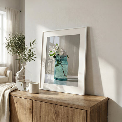 Framed fine art print of white apple blossoms in a vintage blue mason jar on a wooden cabinet.