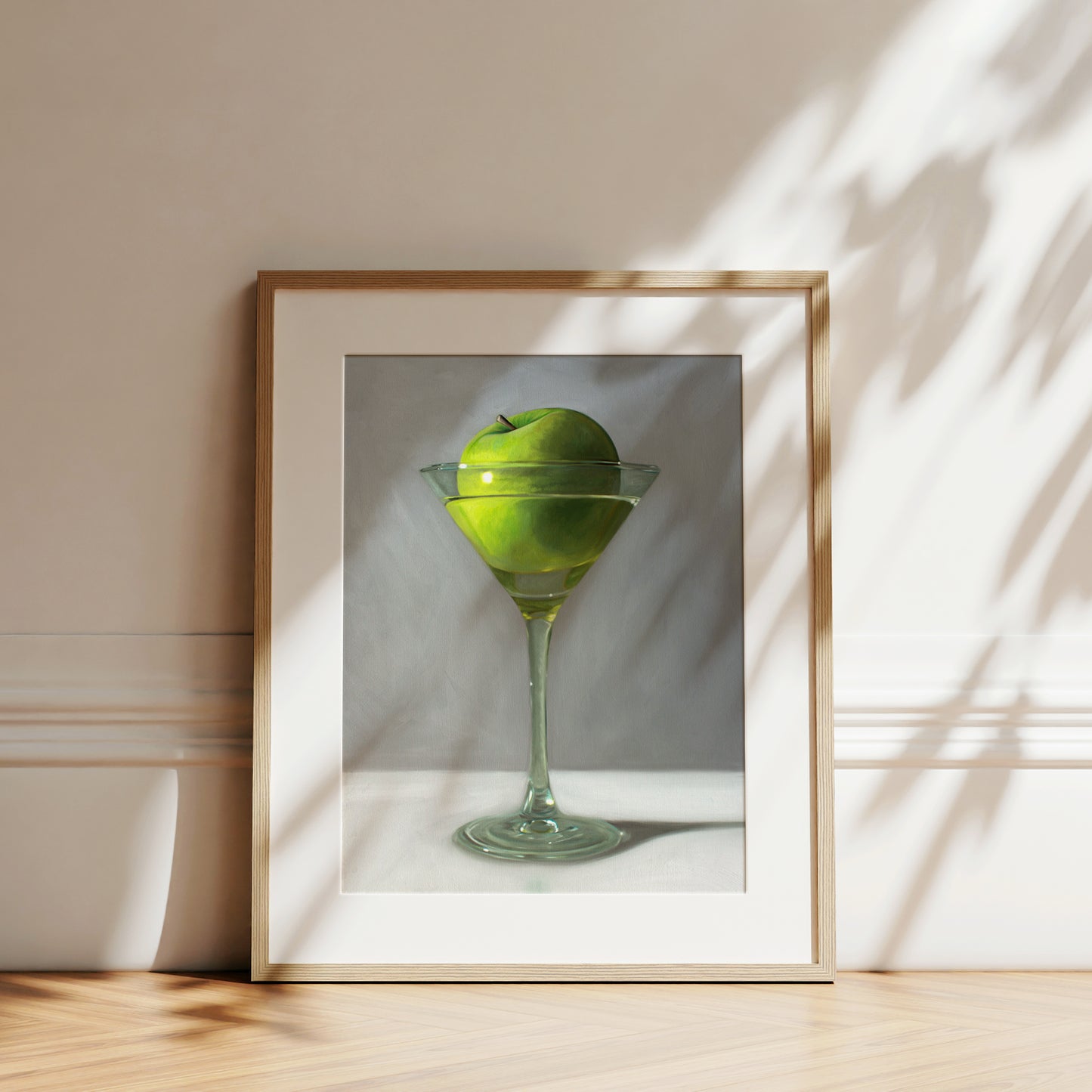 Framed art print of a vibrant green apple in a martini glass, set against a white wall with sun shadows.
