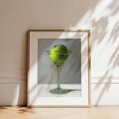 Framed art print of a vibrant green apple in a martini glass, set against a white wall with sun shadows.