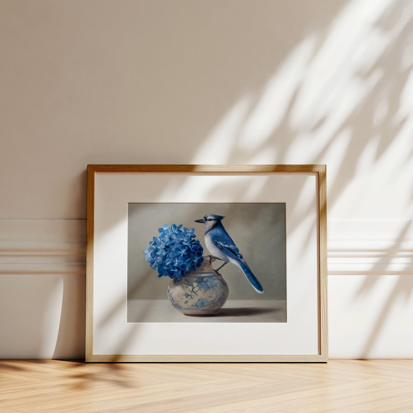 Fine art print of a blue jay perched on a decorative vase with blue hydrangeas, framed in light wood.