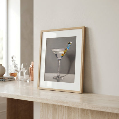 Framed "Bumble Bee Martini" fine art print with olives and a bee stirrer, displayed on a light wood surface.
