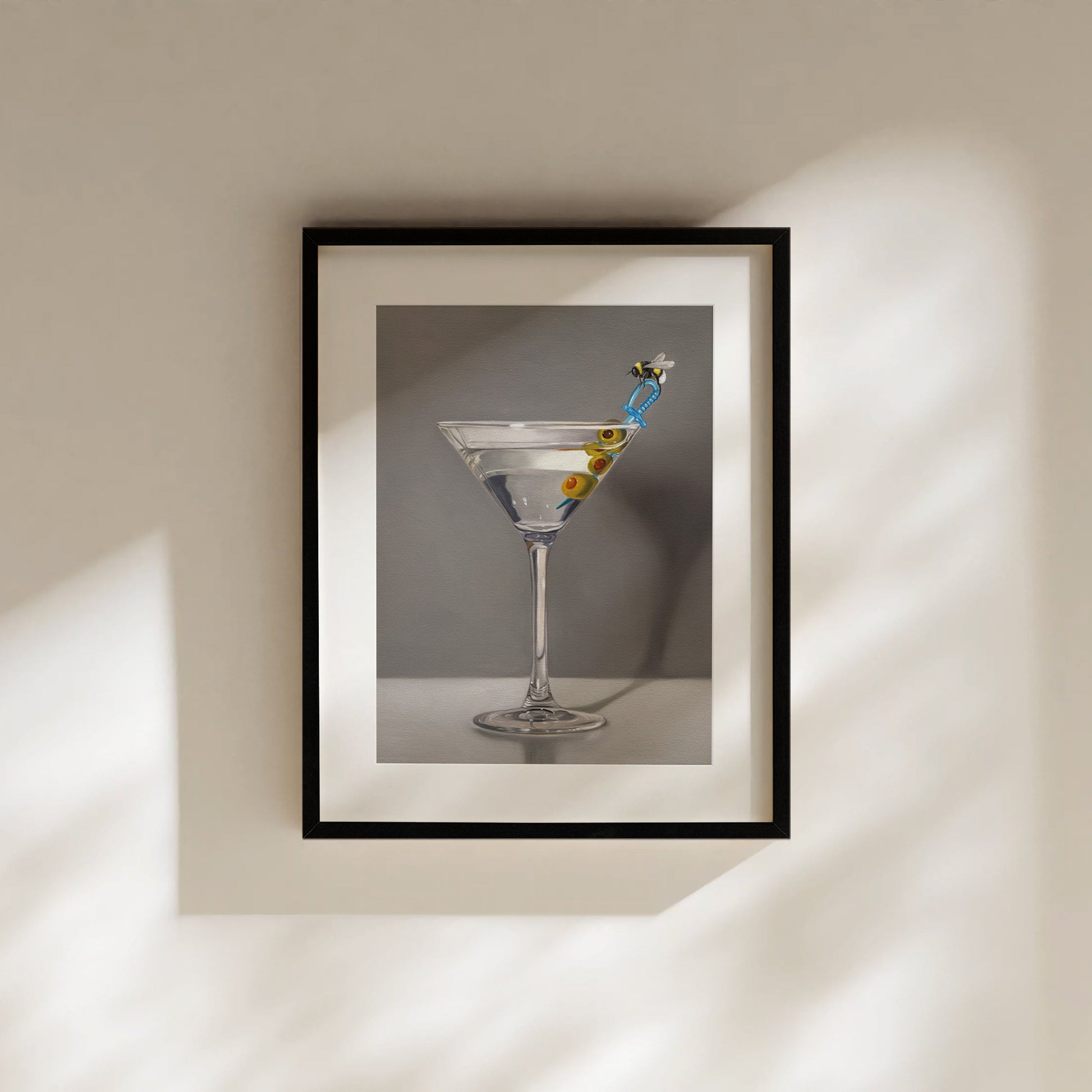 Framed fine art print of a martini with olives and a bumble bee stirrer, on a light wall.