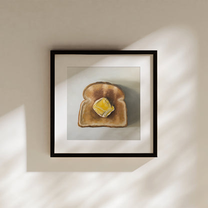 Framed art print of a golden-brown toast slice with melting yellow butter, mounted on a light wall.