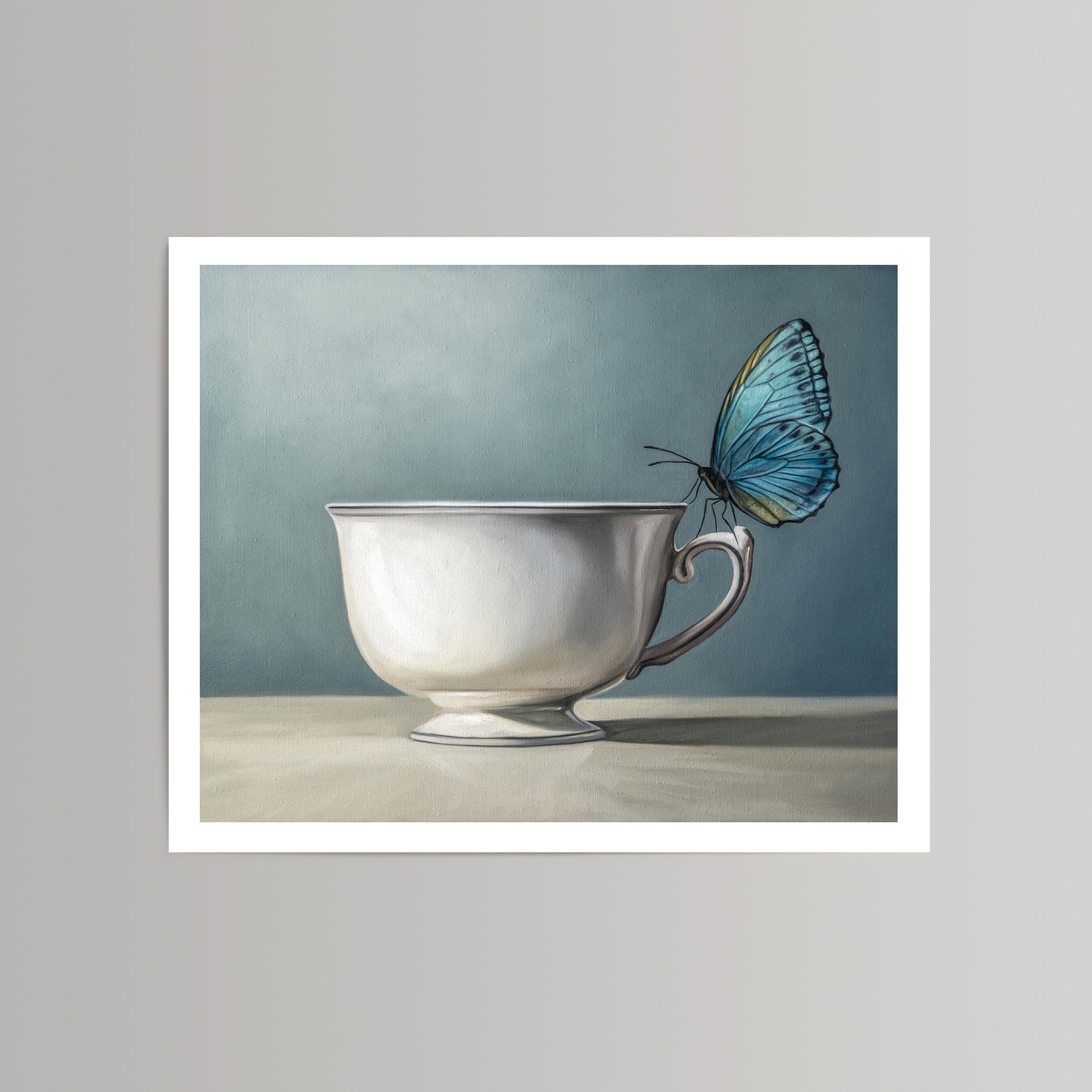 Fine art print of a vibrant blue butterfly perched on a white teacup against a soft blue background.