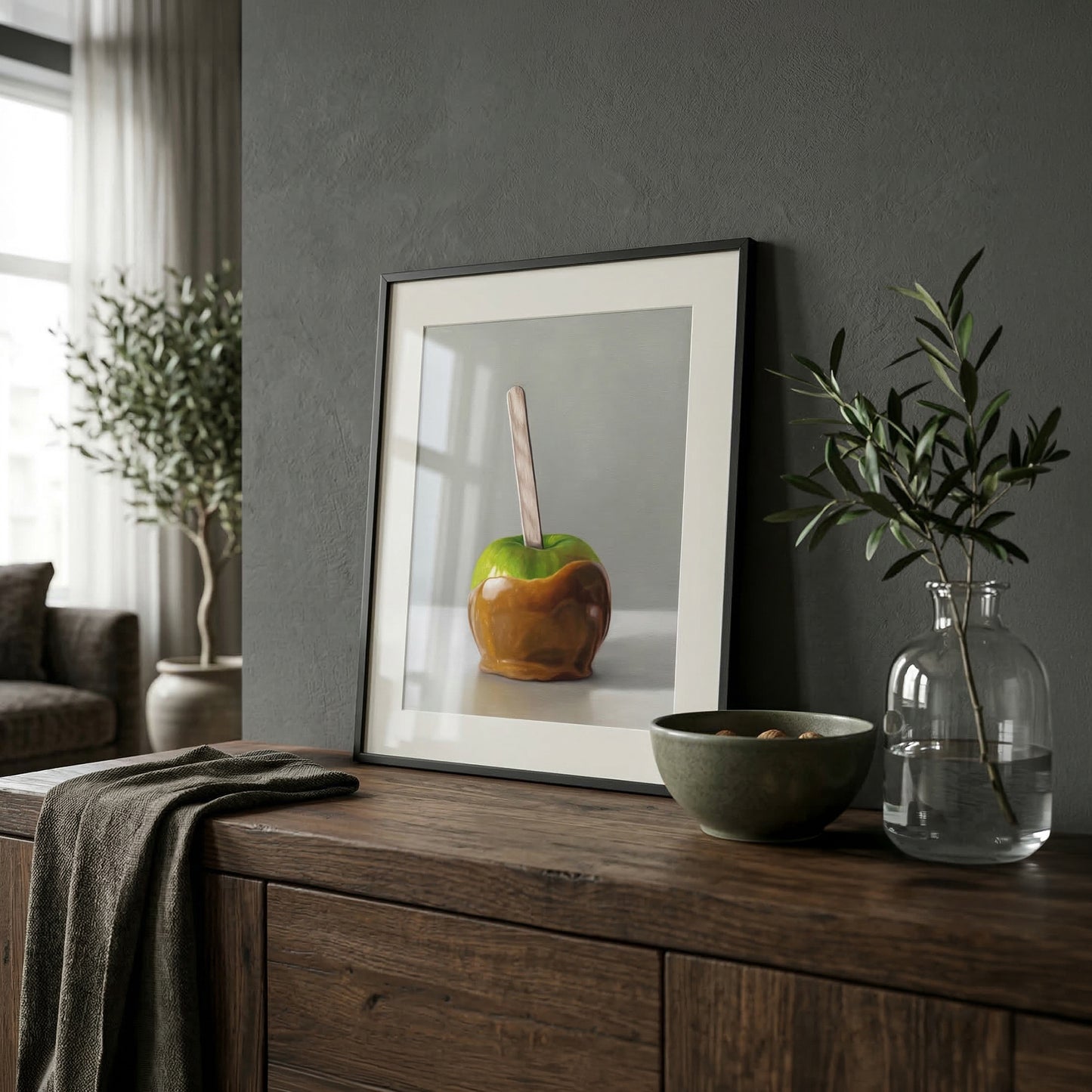 Framed fine art print of a green caramel apple with a stick, displayed on a dark wood cabinet.