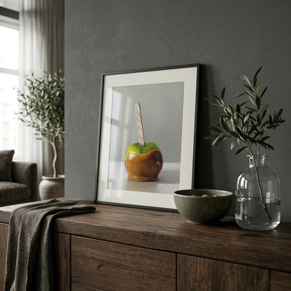 Framed fine art print of a green caramel apple with a stick, displayed on a dark wood cabinet.