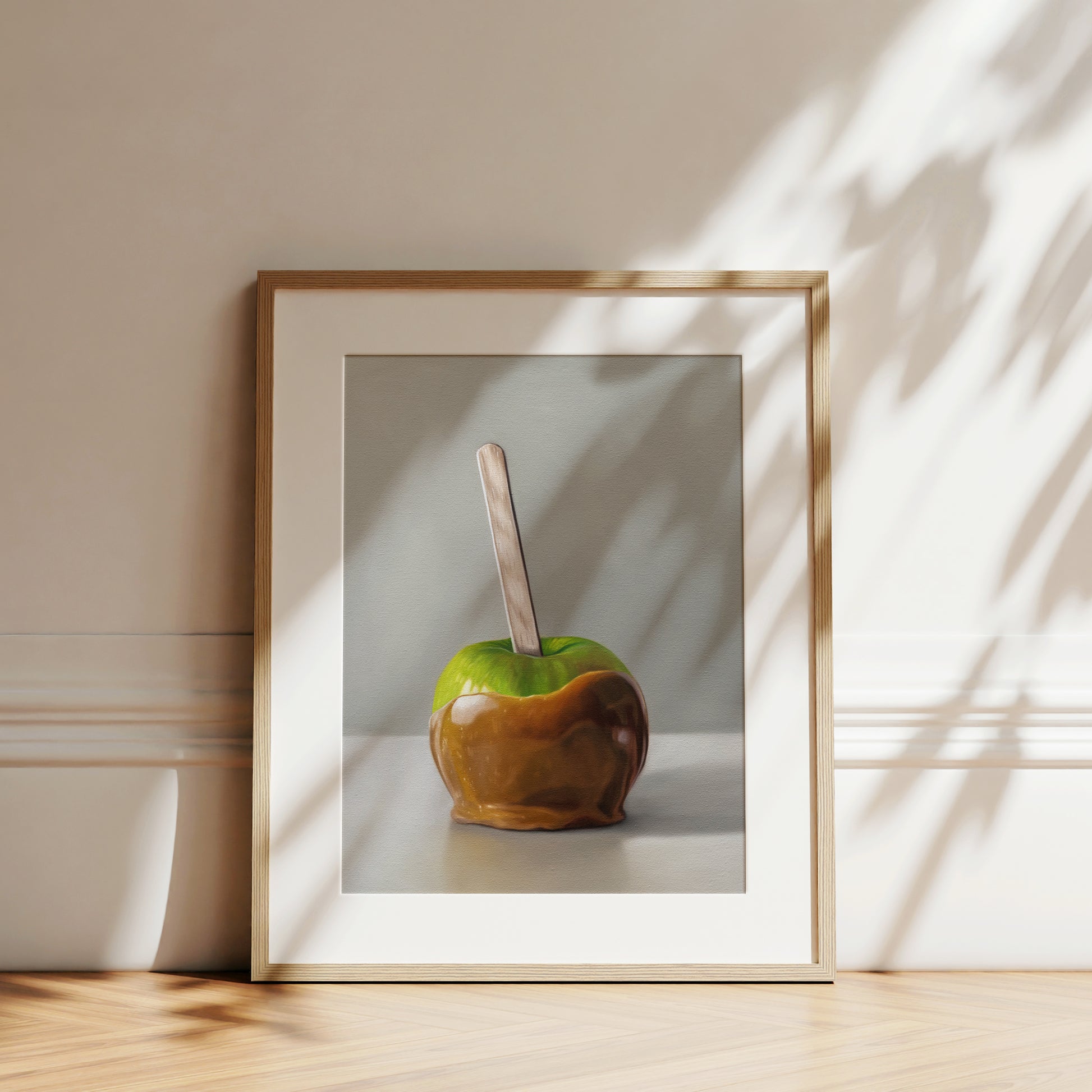 Framed fine art print of a vibrant green caramel apple with a wooden stick, leaning against a white wall.