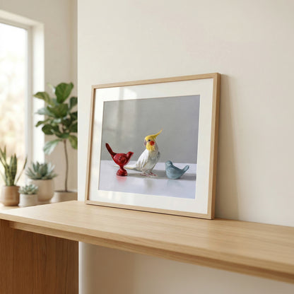Framed fine art print of a cockatiel between red and blue bird figurines on a light wood shelf.
