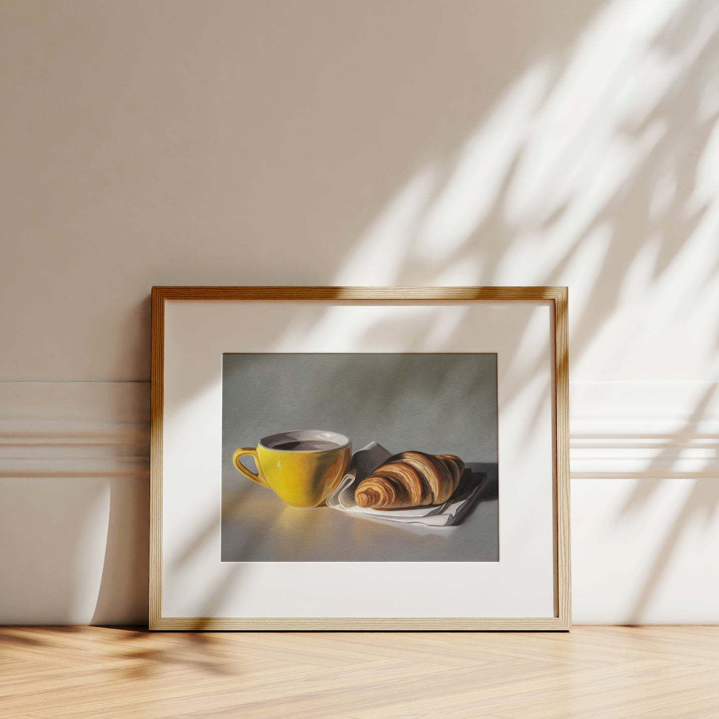 Fine art print of a yellow coffee cup and croissant on a white napkin, framed in light wood.
