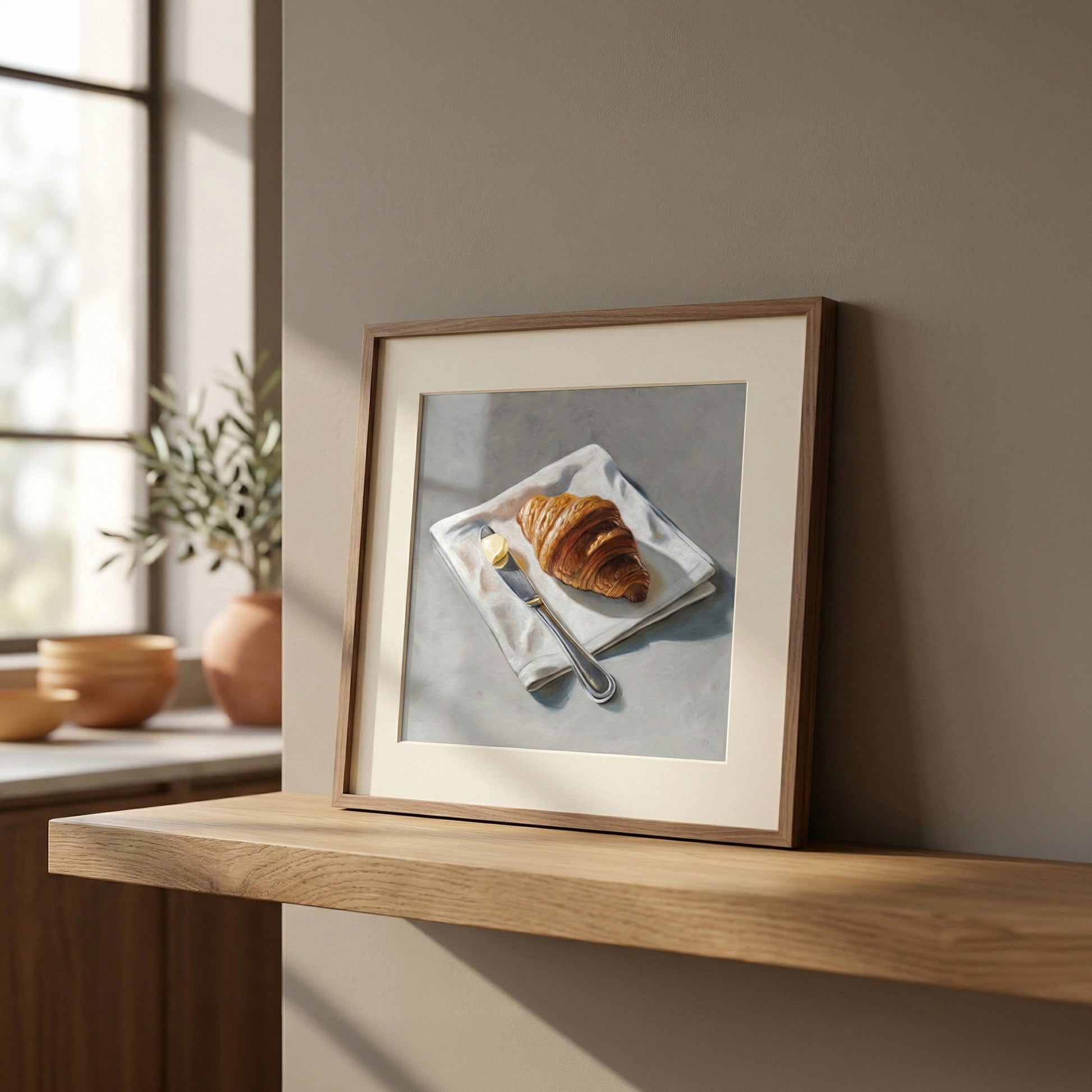 Framed fine art print of a golden croissant, butter, and knife on a white napkin, displayed on a wooden shelf.