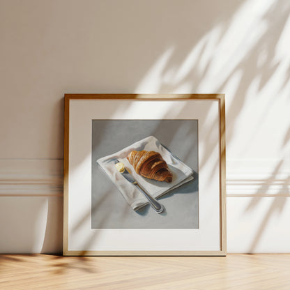 Framed fine art print of a golden croissant, butter, and knife on a white napkin, with sunlit shadows.