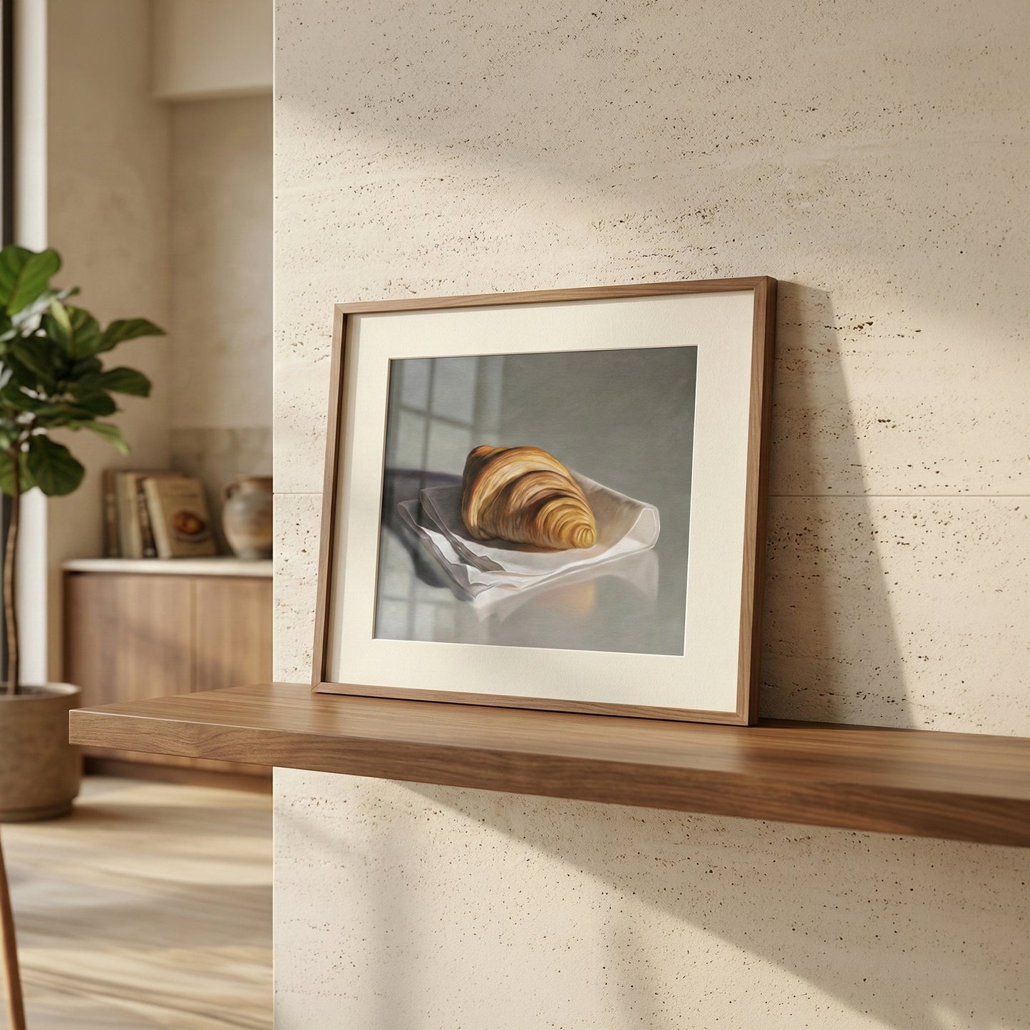 Framed fine art print of a golden croissant on white paper, displayed on a wooden shelf in a modern room.