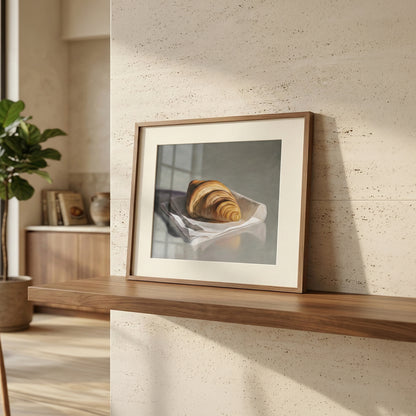 Framed fine art print of a golden croissant on white paper, displayed on a wooden shelf in a modern room.