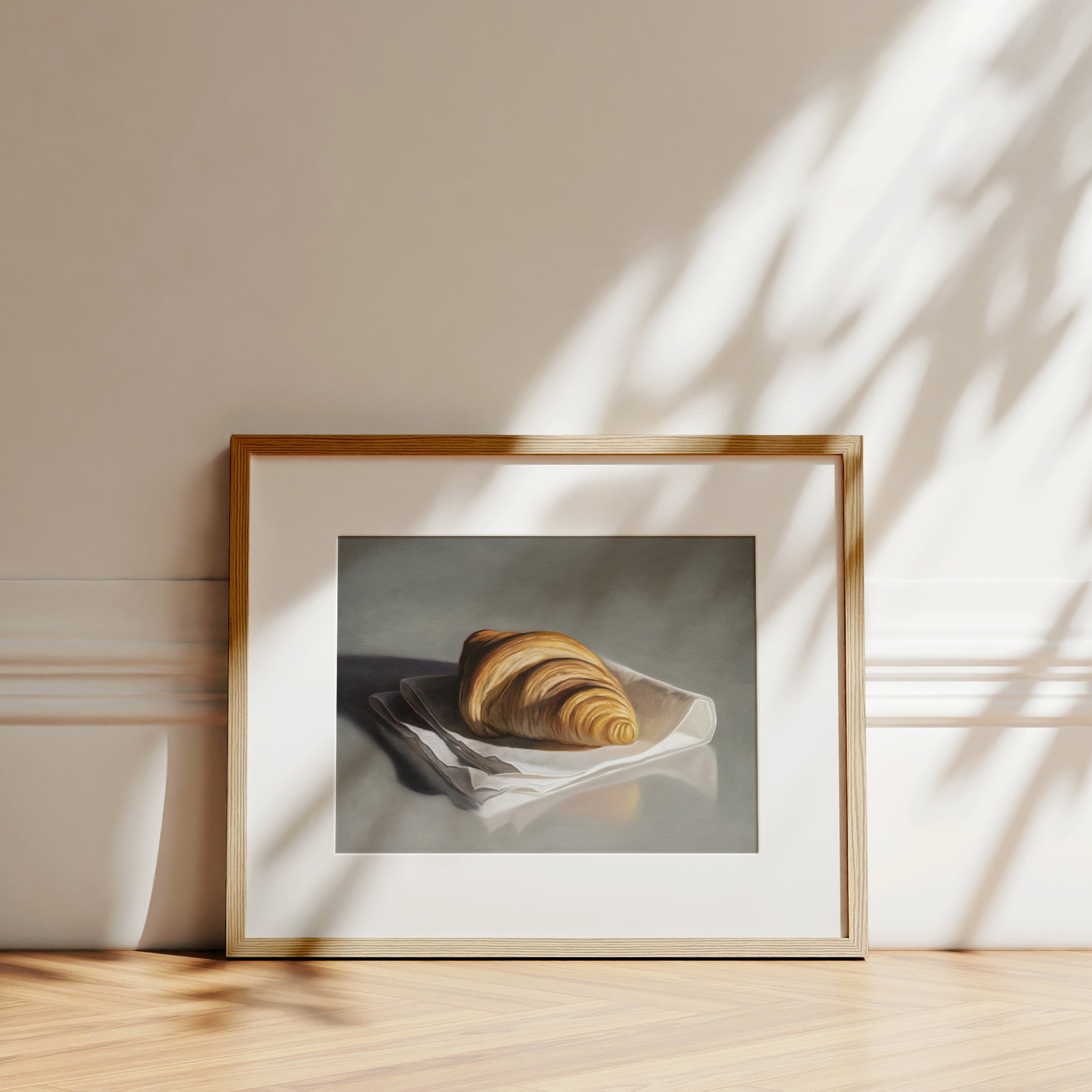Framed fine art print of a golden croissant on a white napkin, set against a neutral wall.