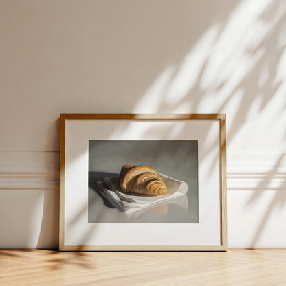 Framed fine art print of a golden croissant on a white napkin, set against a neutral wall.