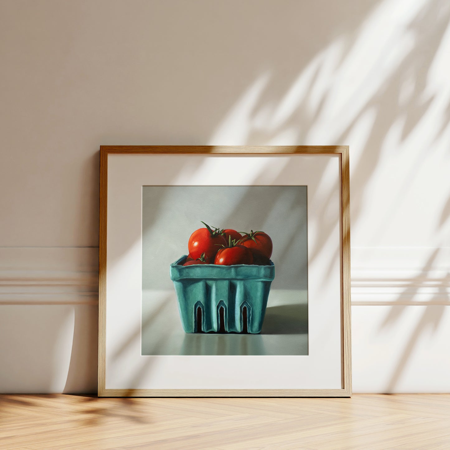 Framed fine art print of red tomatoes in a teal berry basket, with a natural wood frame and white mat.