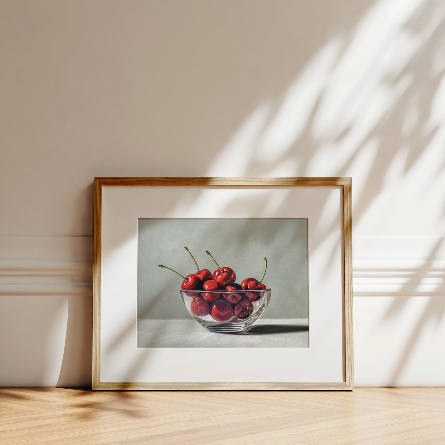 Fine art print of a glass bowl filled with vibrant red cherries, framed in light wood with a white mat.