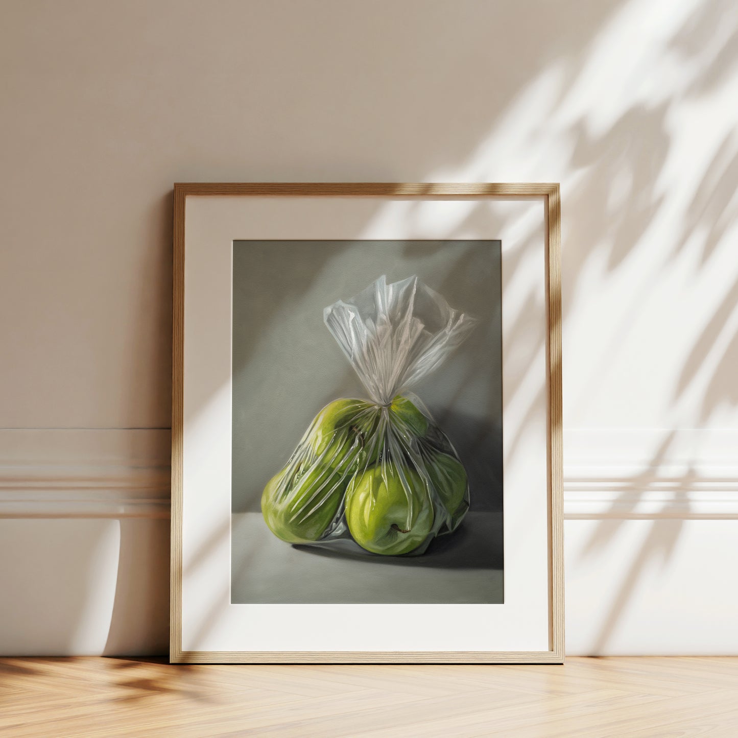 Framed fine art print of three green Granny Smith apples in a clear plastic bag, hanging on a white wall.