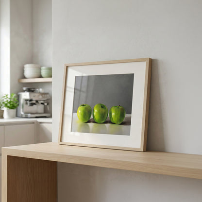 Fine art print of three vibrant green apples in a light wood frame, displayed on a modern wooden shelf.