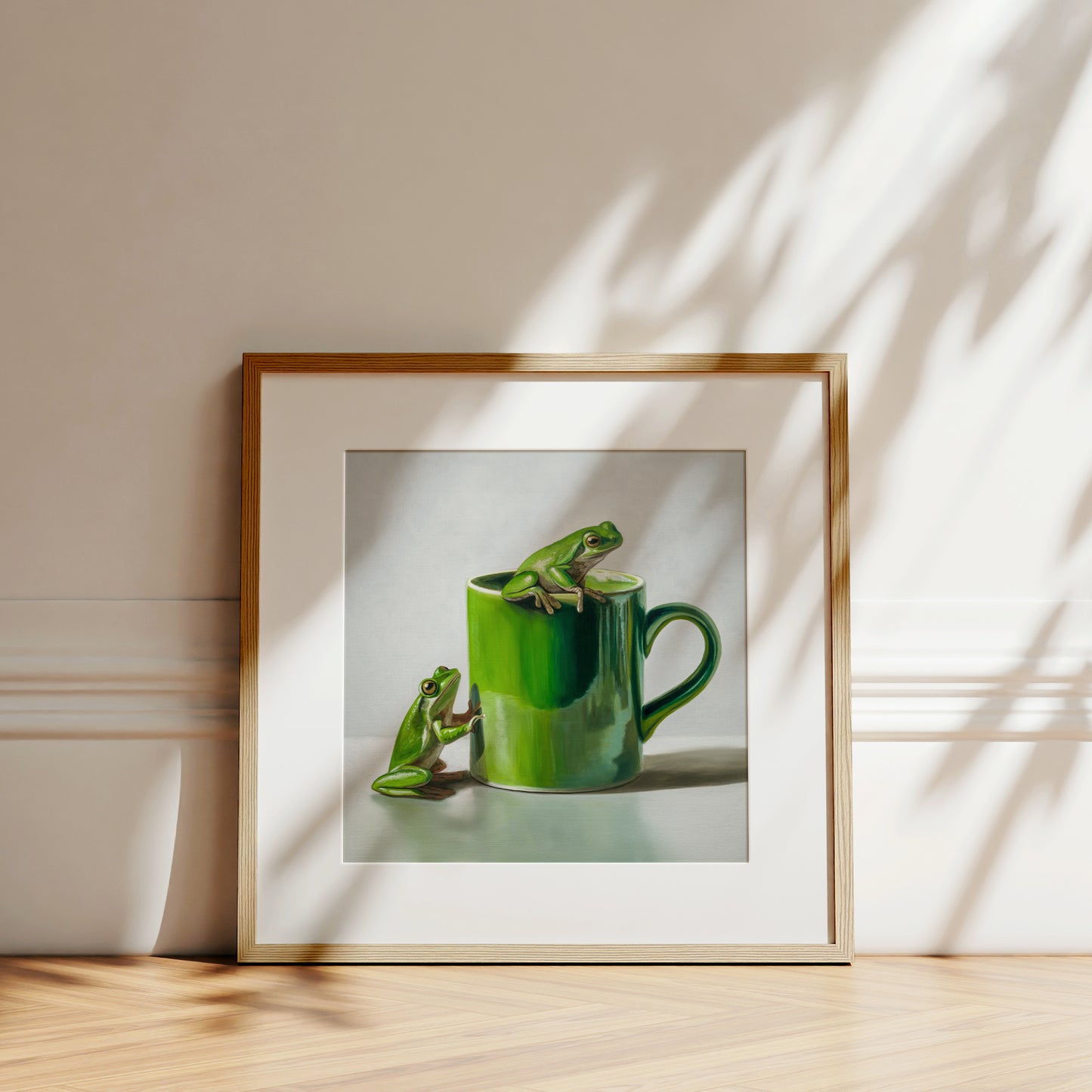 Fine art print of two green frogs, one on a vibrant green mug, in a natural wood frame.