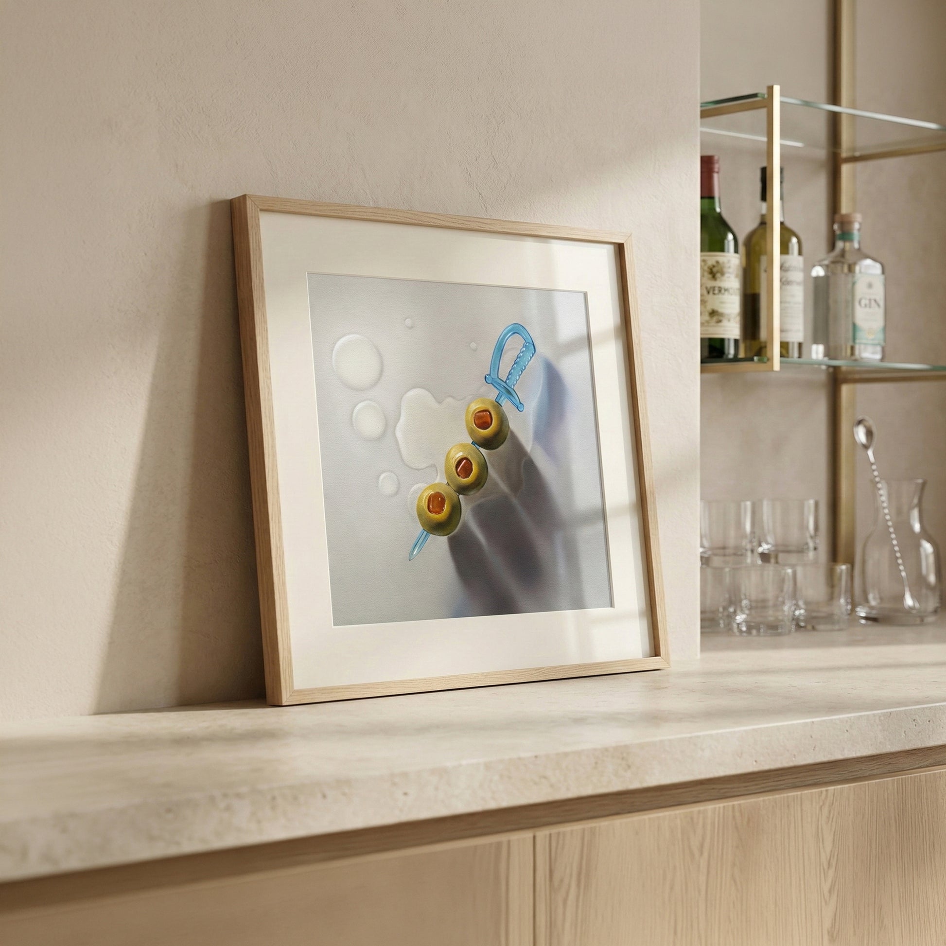 Framed art print of three green olives on a blue sword pick, with water droplets, on a light countertop.