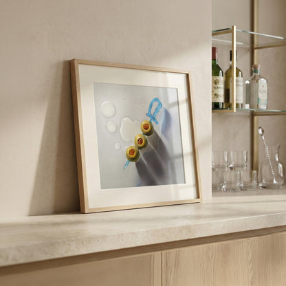 Framed art print of three green olives on a blue sword pick, with water droplets, on a light countertop.