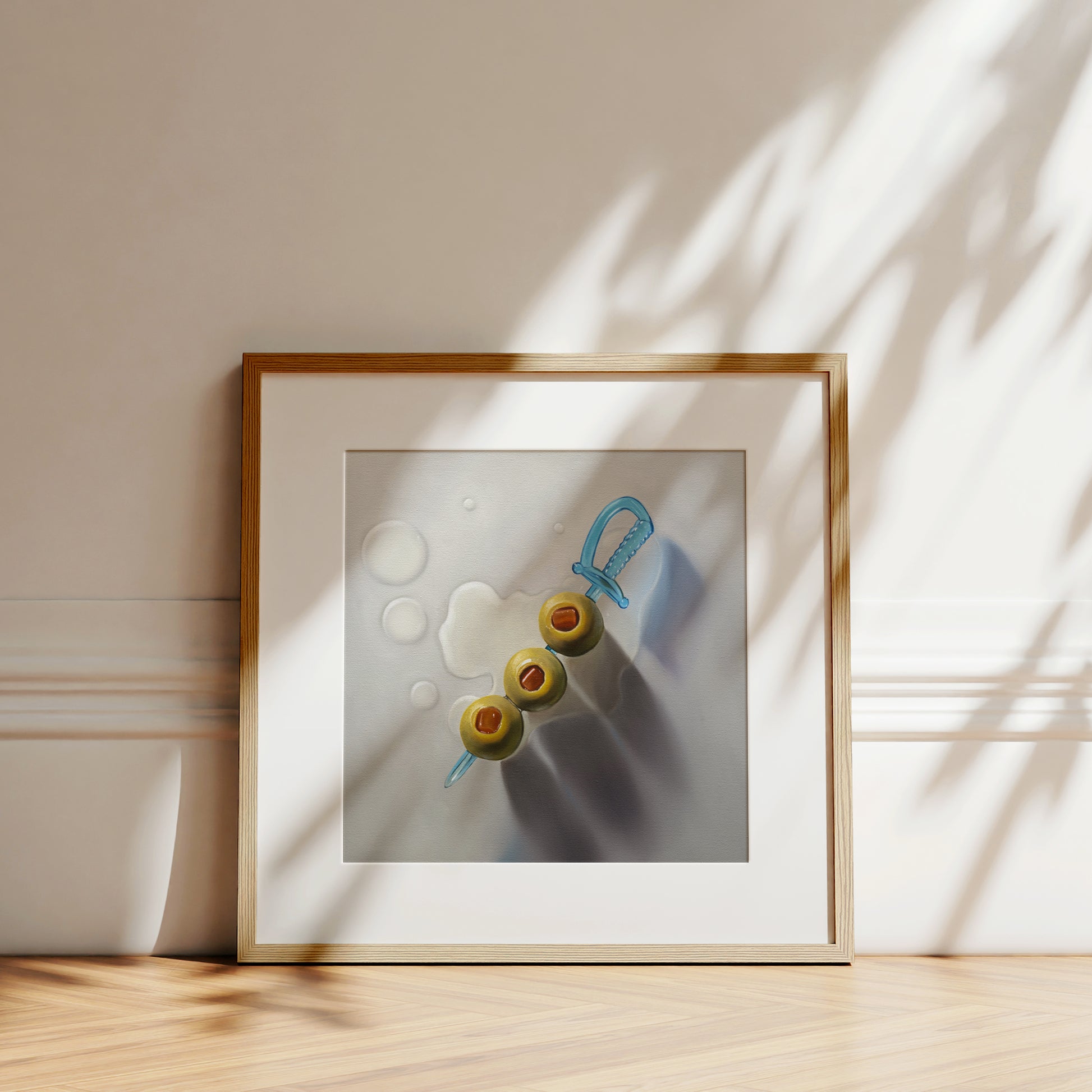 Framed art print of three green olives on a blue sword skewer with water drops, leaning on a wall.