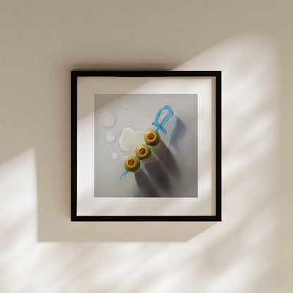 Framed art print of three green olives on a blue sword skewer, with subtle liquid rings on a light background.