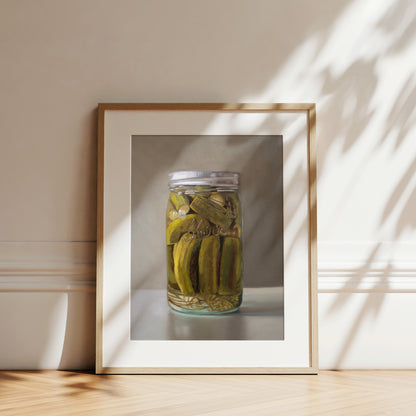 Framed fine art print of a glass jar filled with green homemade pickles, on a light wall.