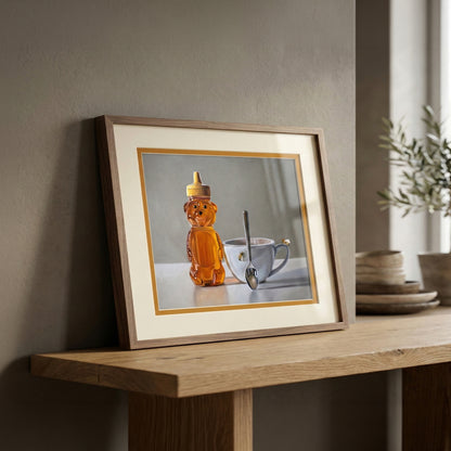 Framed fine art print of a honey bear bottle, white teacup with a bee, and spoon on a light background.