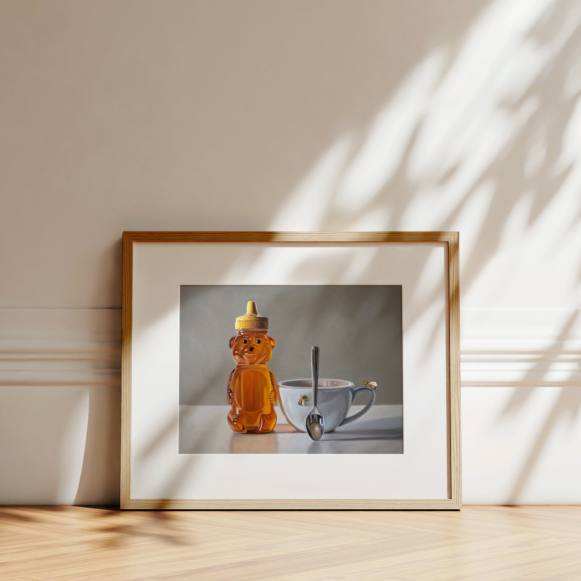 Framed fine art print of a honey bear bottle, a teacup with a bee, and a spoon, on a light wood surface.