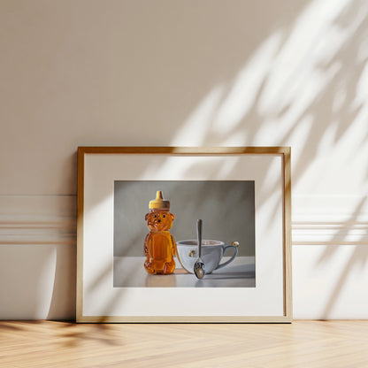 Framed fine art print of a honey bear bottle, a teacup with a bee, and a spoon, on a light wood surface.