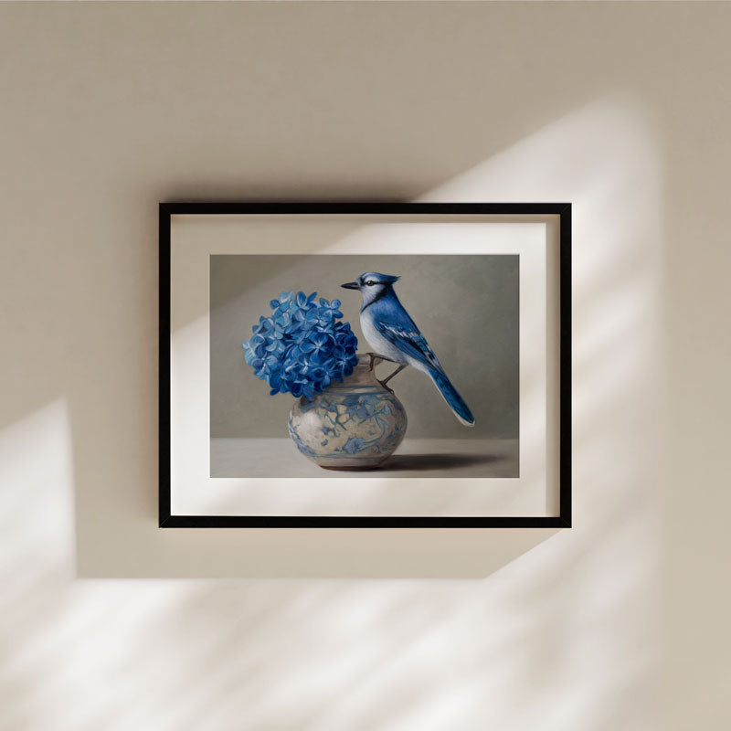 Fine art print of a blue jay perched on a vase with blue hydrangeas, displayed in a black frame on a neutral wall.