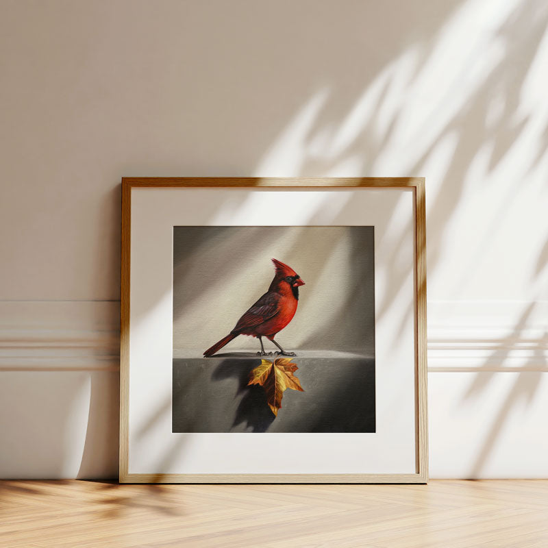 Fine art print of a red cardinal perched above a golden maple leaf, framed in light wood on a white wall.