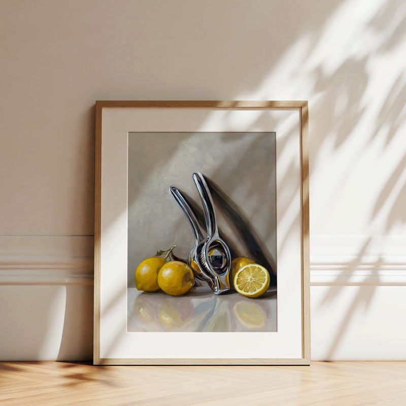 Fine art print of a chrome citrus press and yellow lemons in a light wood frame, displayed against a neutral wall.
