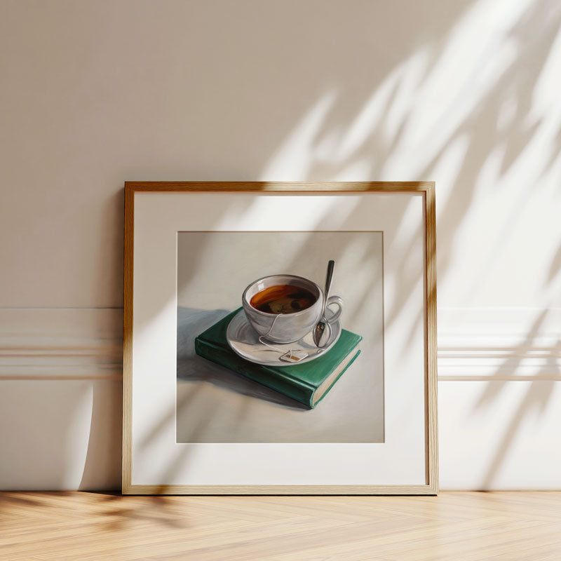 Fine art print of a tea cup on a green book in a light wood frame, displayed against a wall with soft leaf shadows.