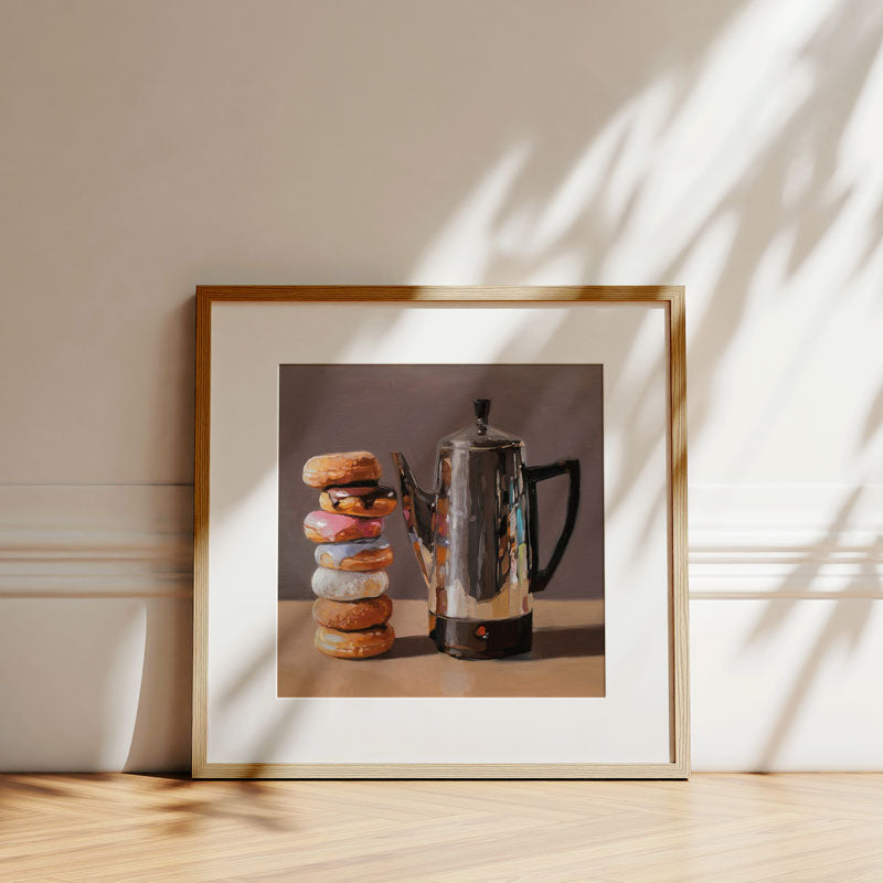 Fine art print of a stacked donut tower and silver percolator in a light wood frame against a neutral wall.