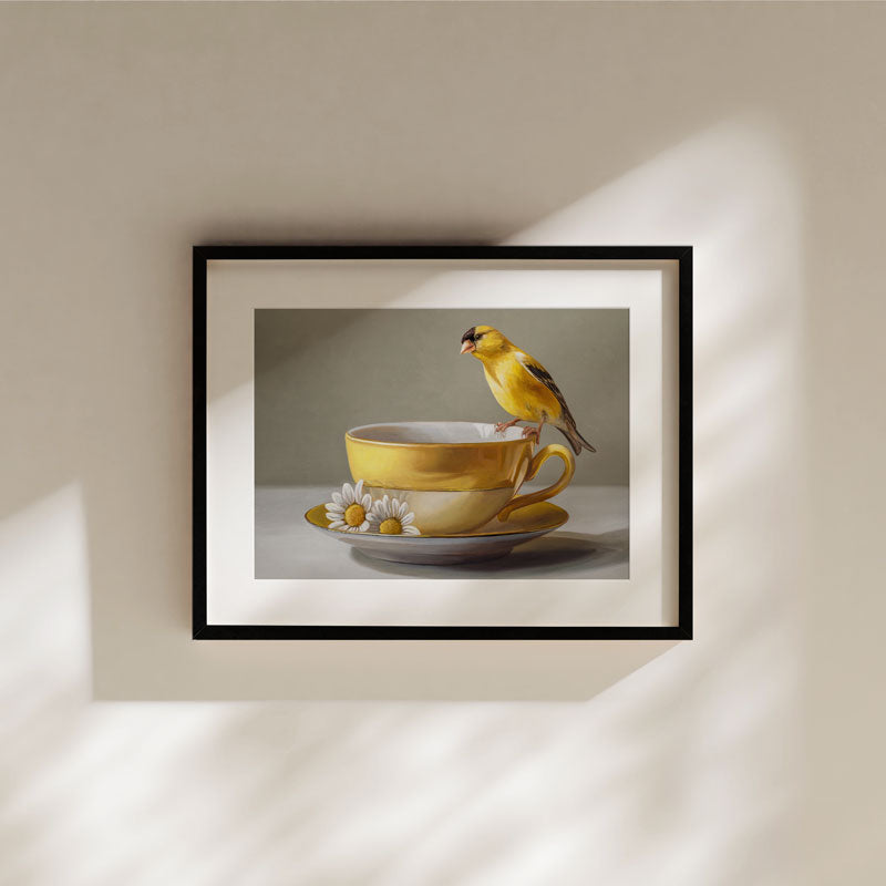 Fine art print of a yellow goldfinch perched on a yellow teacup with daisies, framed in black on a neutral wall.