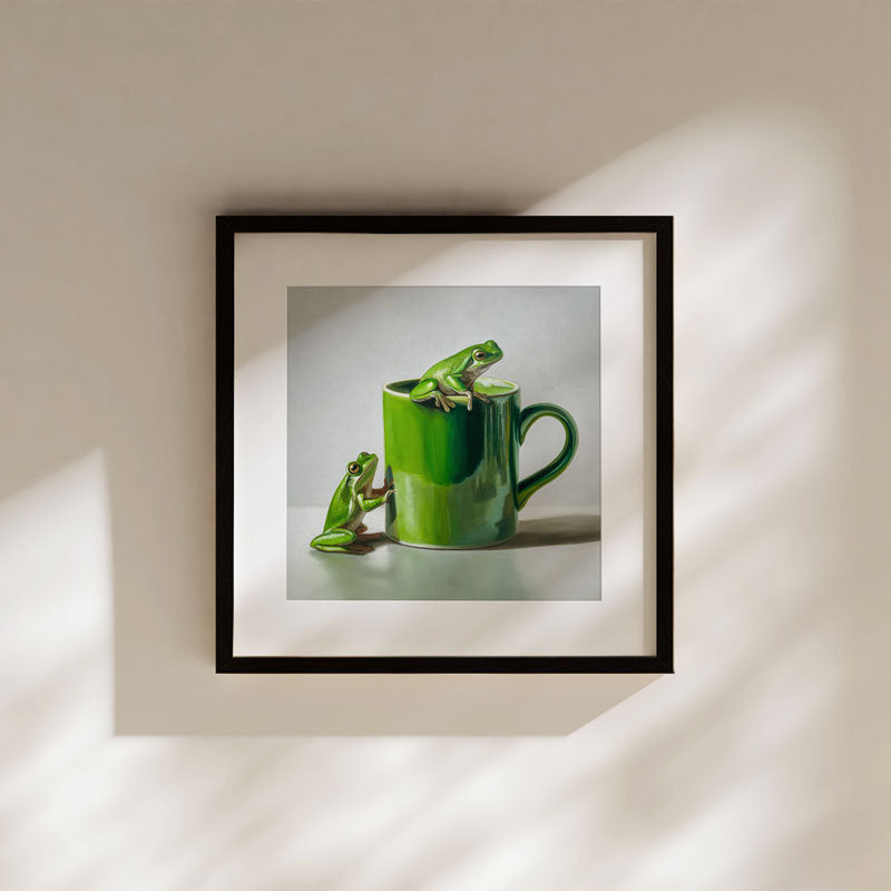 Fine art print of two green frogs on a green coffee mug, displayed in a black frame on a neutral wall.