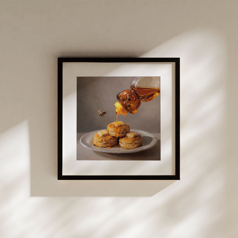 Fine art print of honey pouring from a bear bottle onto buttered biscuits, framed in black on a neutral wall.
