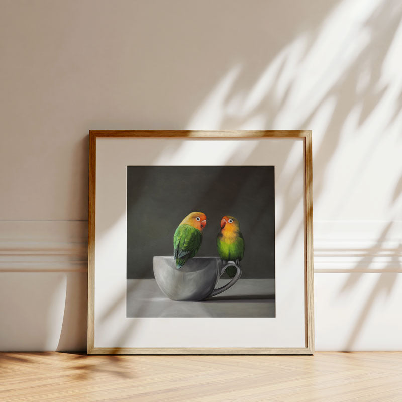 Fine art print of two green lovebirds perched on a porcelain cup, framed in a light wood frame against a white wall.