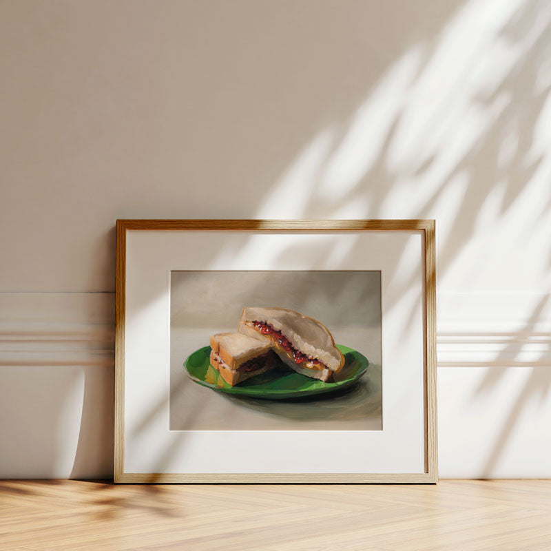 Fine art print of a peanut butter and jelly sandwich on a green plate, displayed in a light wood frame on a floor.