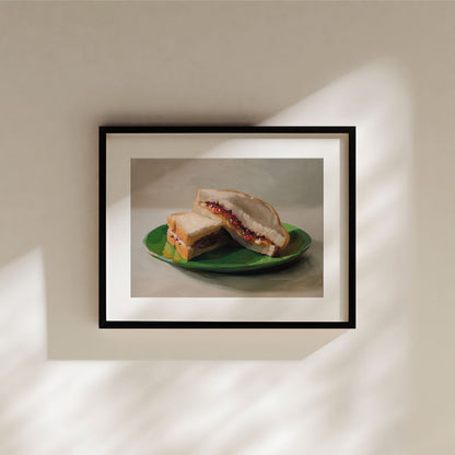 Fine art print of a peanut butter and jelly sandwich on a green plate, framed in black with a white mat border.
